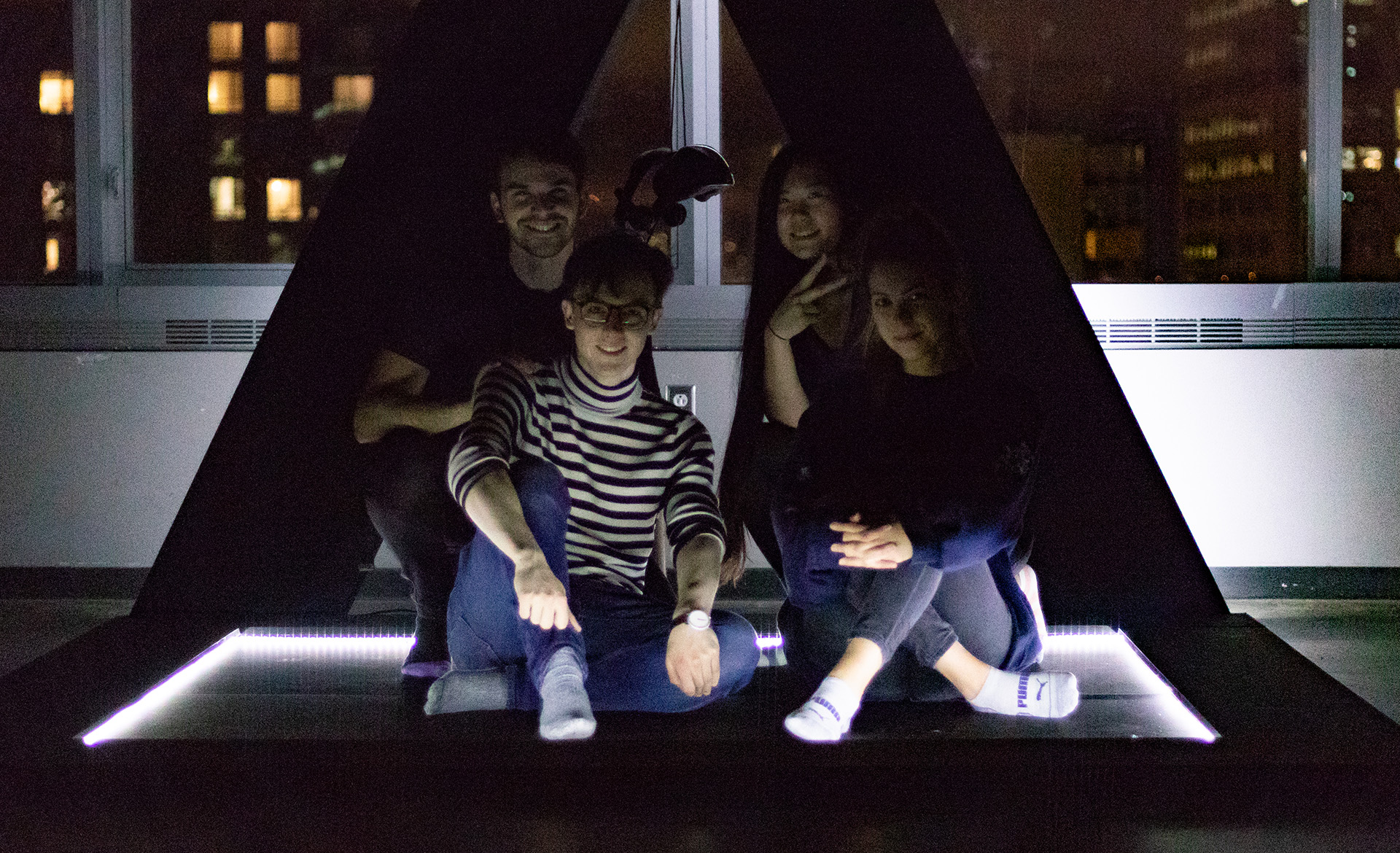 Photograph of an installation sculpture shaped like a triangle in a dark room and 4 people sitting in front of it, smiling.